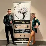 Un danseur et une danseuse en costumes de scène verts, de part et d'autre du roll-up Villa Vida.